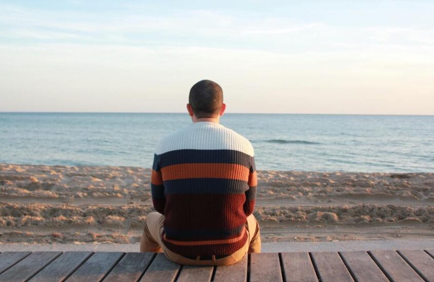 Man sitting on wooden panel facing the ocean.